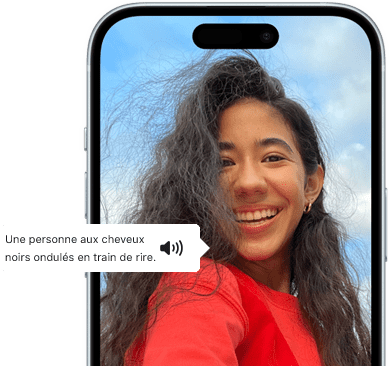 Un iPhone 15 montrant la fonctionnalité VoiceOver énonçant les informations figurant sur l’image : Une personne aux cheveux noirs ondulés en train de rire
